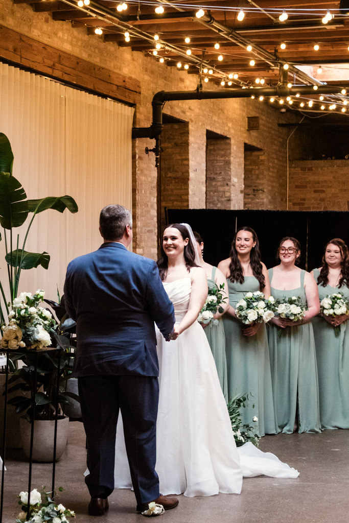 Bride and groom hold hands during an indoor wedding ceremony at The Arbory, with bridesmaids in green dresses behind them