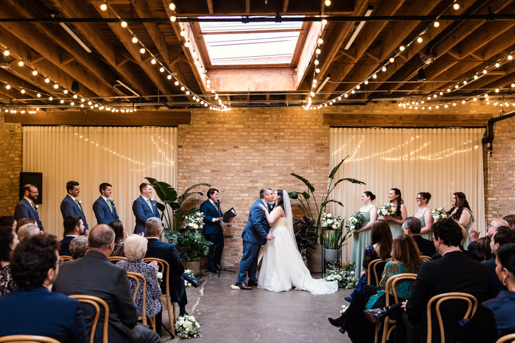 Bride and groom kiss at their indoor wedding ceremony at The Arbory, surrounded by guests