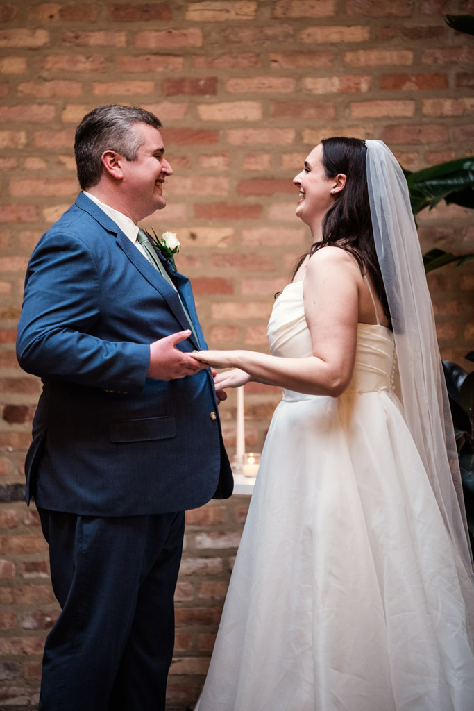 Bride and groom smiling and holding hands during their wedding ceremony in front of a brick wall at The Arbory