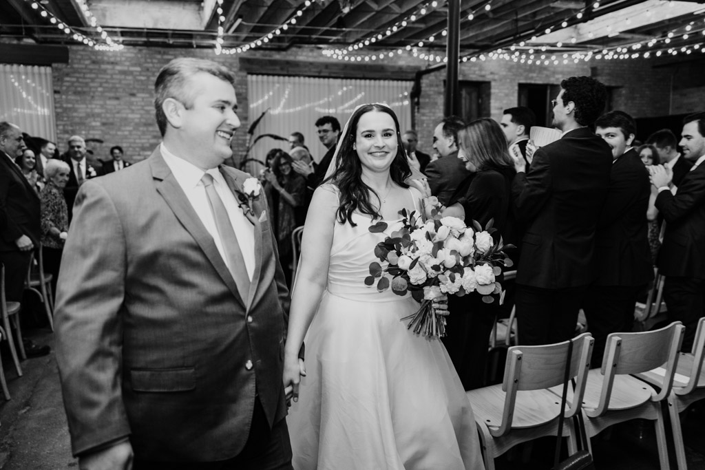 Black and white photo of bride and groom smiling and holding hands, walking down the aisle after their wedding ceremony at The Arbory