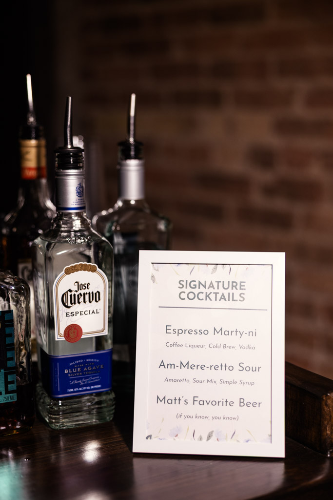Bottles of liquor next to a menu listing signature cocktails, including Espresso Marty-ni and Am-Mere-retro Sour for wedding reception at The Arbory
