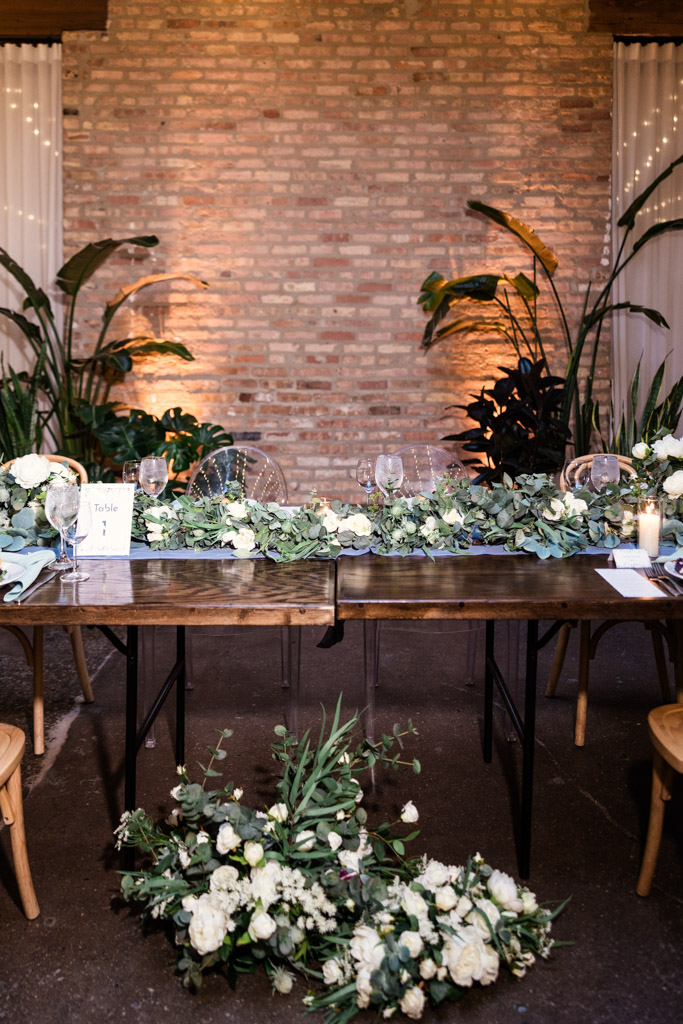 Wedding head table at The Arbory with greenery, white flowers, candles, and a brick wall in the background