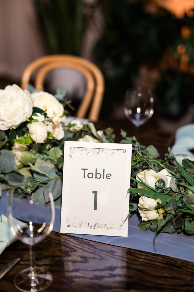 Table marker on a wooden guest table with white flowers, greenery, and wine glasses for wedding reception at The Arbory