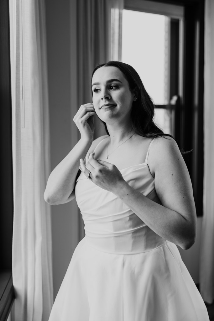 Black and white photo of bride in a white dress putting on an earring while standing by a window at The Robey Chicago