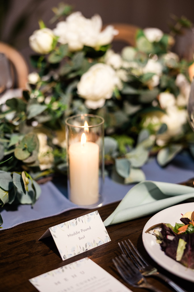 A lit candle, flowers, and a name card on a guest table set with a salad plate and menu for wedding reception at The Arbory
