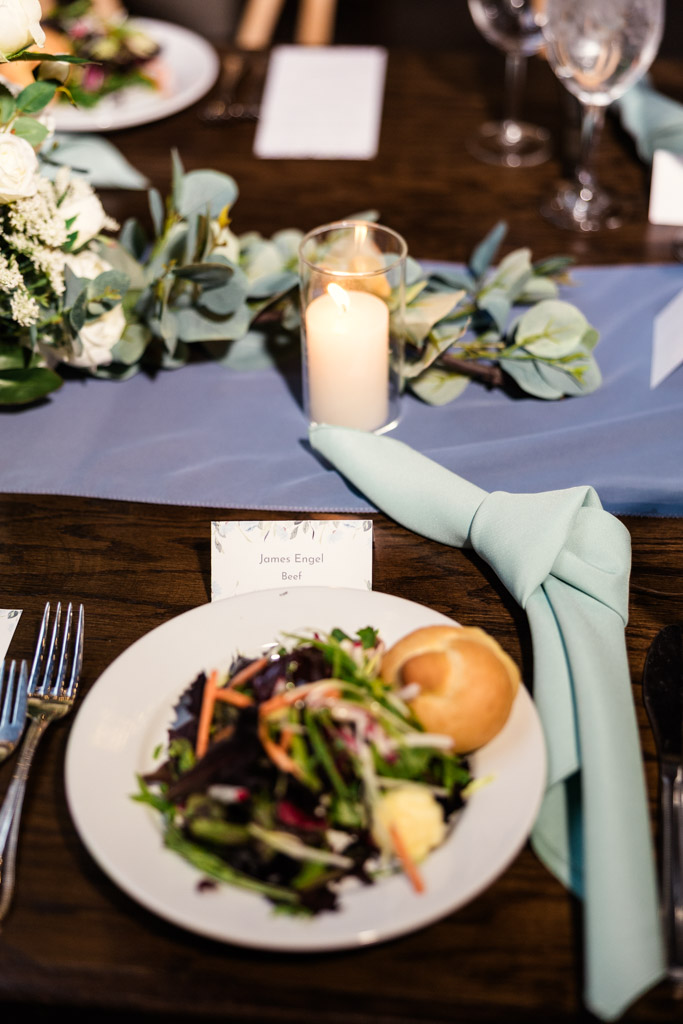 Guest place setting with salad and a roll on a table set with a candle, napkin, utensils, and a name card for wedding reception at The Arbory