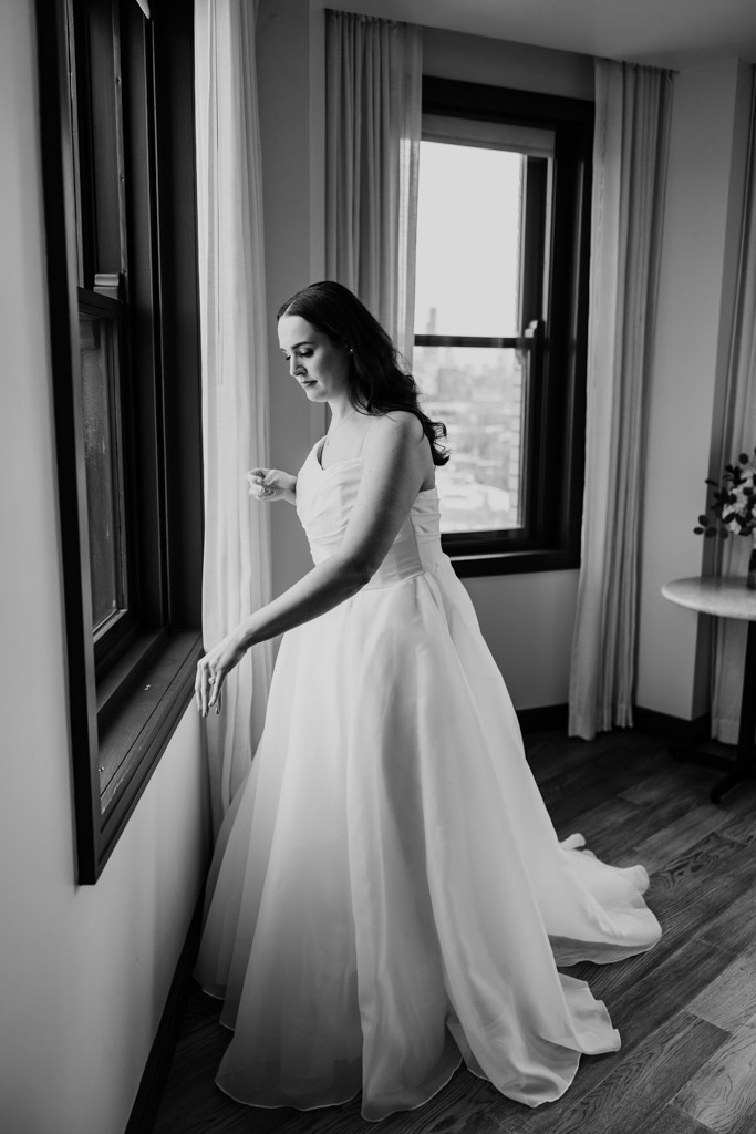 Black and white photo of bride in a wedding dress looking out the window while getting ready at The Robey Chicago