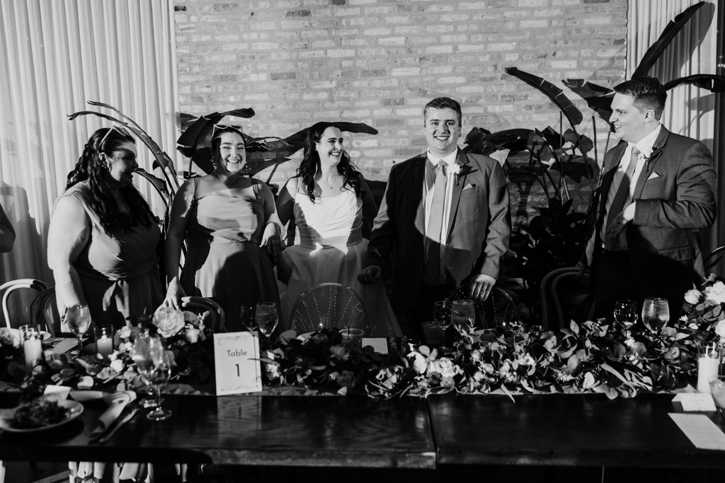Black and white photo of bride, groom and wedding party members standing smiling behind a decorated table at wedding reception at The Arbory.