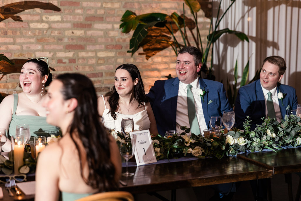 Newlyweds listen to a speech as they sit at the head table, smiling and laughing at their wedding reception held at The Arbory