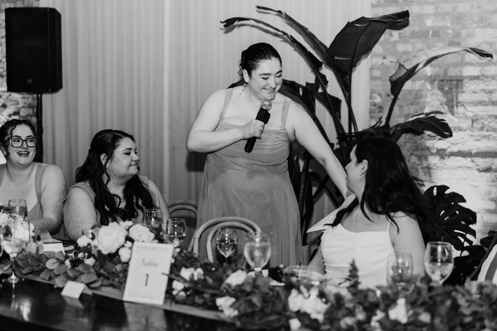 Black and white photo of maid of honor giving a speech with a microphone at a wedding reception at The Arbory, smiling at the bride