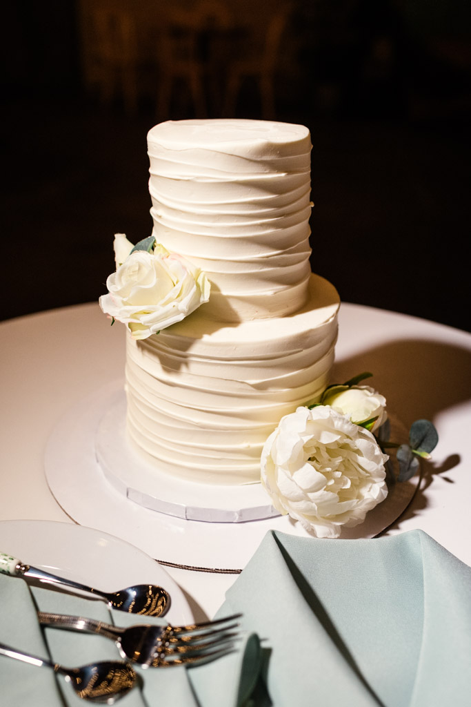 A two-tier white cake with textured icing and white flowers, set on a table with plates and silverware for wedding reception at The Arbory