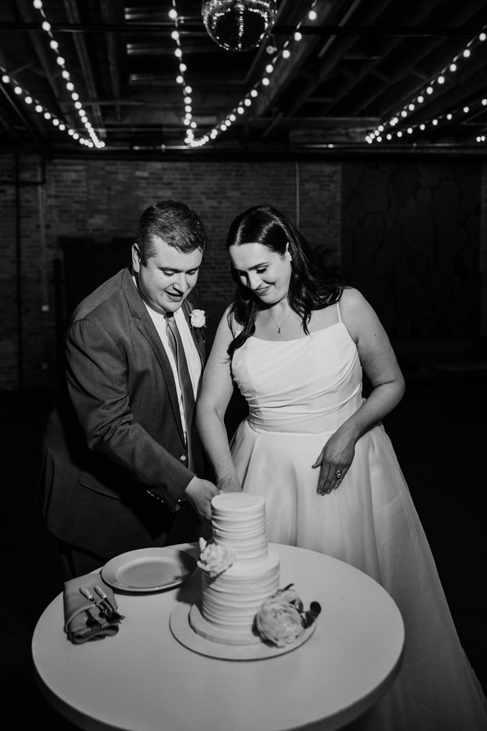 Black and white photo of bride and groom in formal attire cutting their wedding cake together under string lights at The Arbory