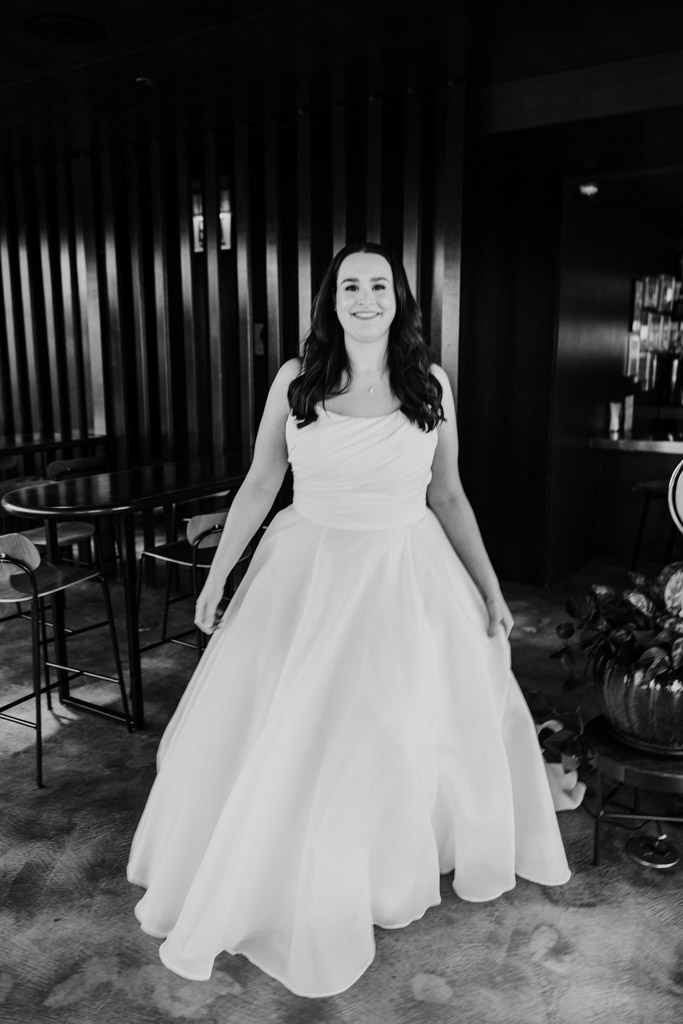 Black and white portrait of bride in a white gown stands inside The Up Room at The Robey Chicago with tables and chairs in the background