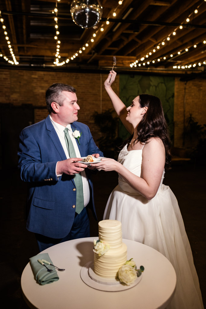 Bride and groom smile, holding forks, ready to eat their wedding cake under string lights at The Arbory
