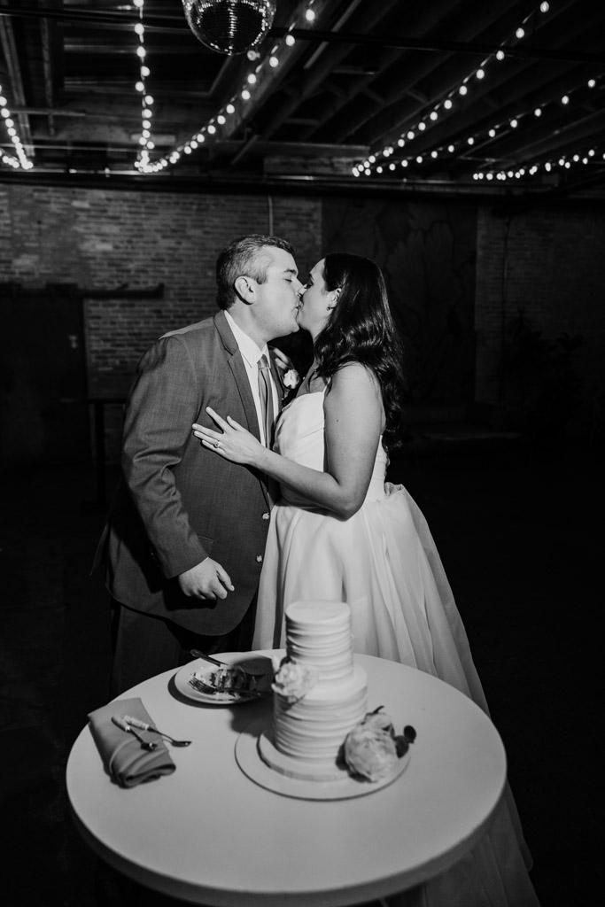 Newlywed couple in wedding attire kisses by a table with a tiered cake under string lights during their wedding reception at The Arbory