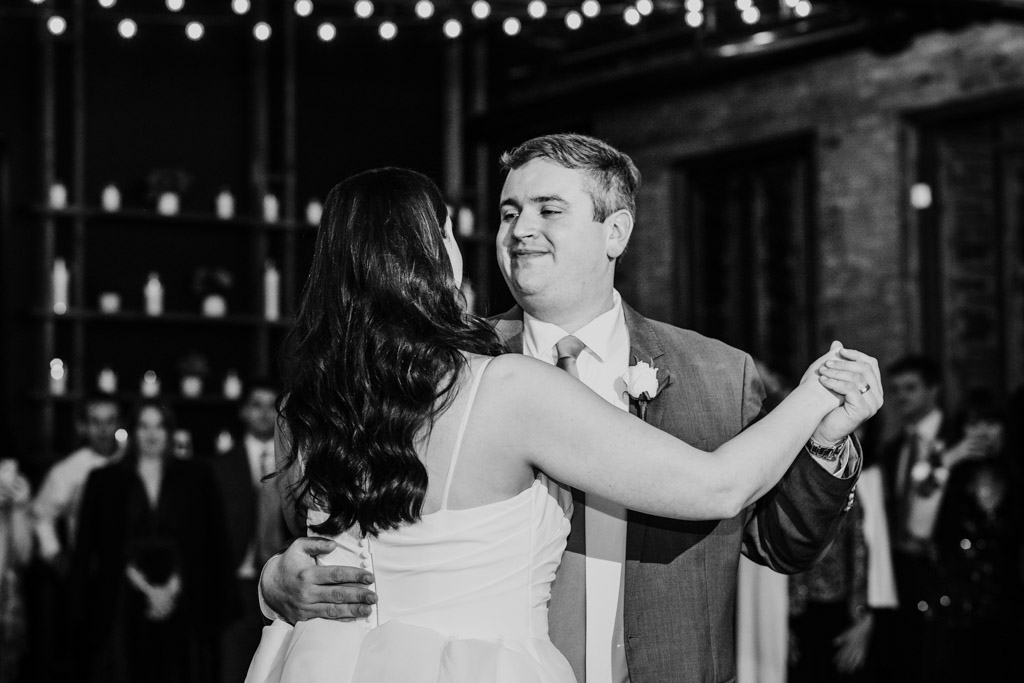 Black and white photo of newlywed couple in formal attire during their first dance under string lights at their wedding reception at The Arbory