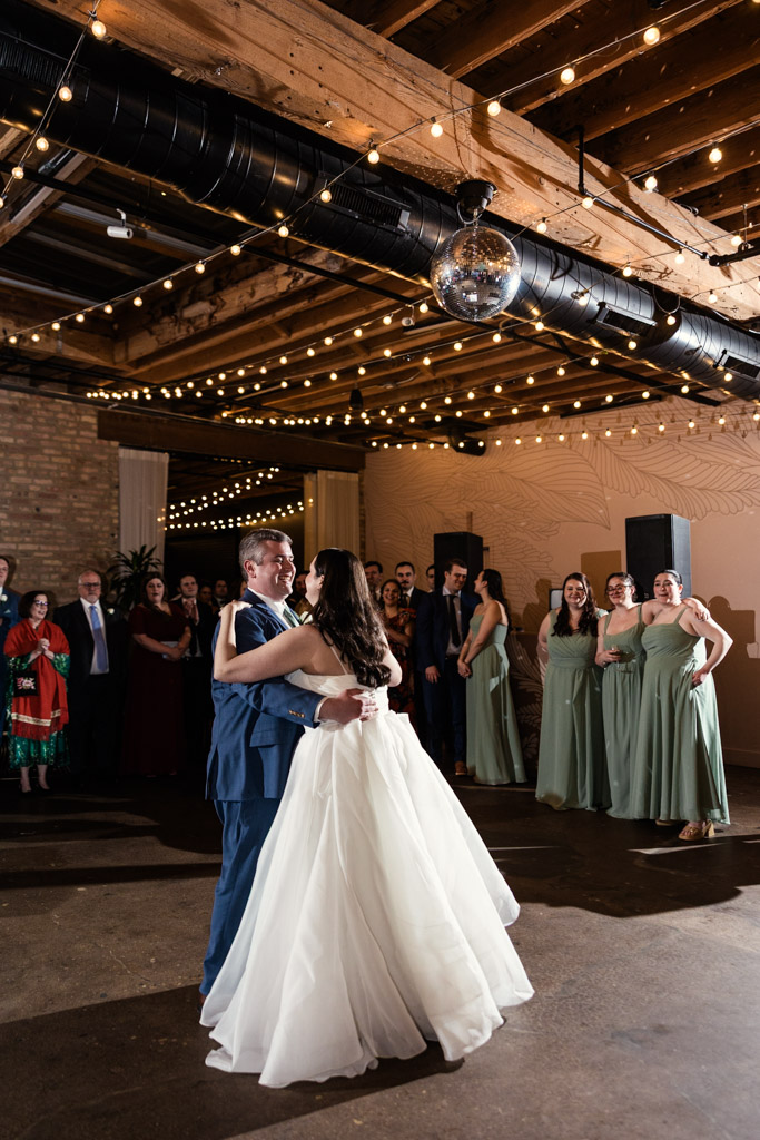 Bride and groom share their first dance under string lights at The Arbory as guests watch