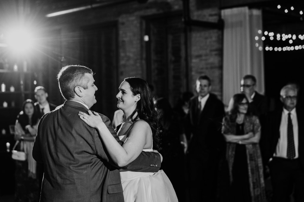 Black and white photo of bride and groom dancing together, surrounded by guests at their wedding reception at The Arbory