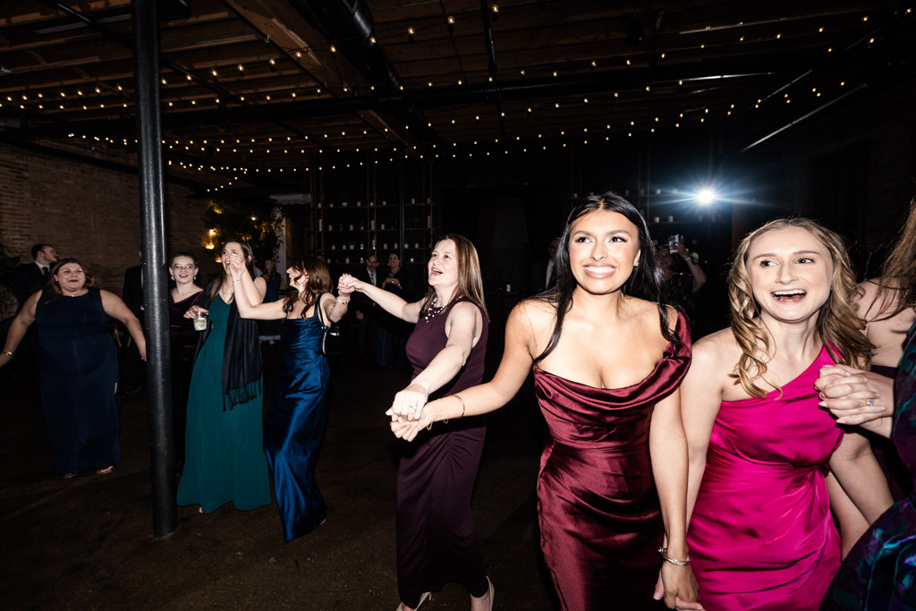Guests in formal dresses laugh and hold hands while dancing under string lights during wedding reception at The Arbory
