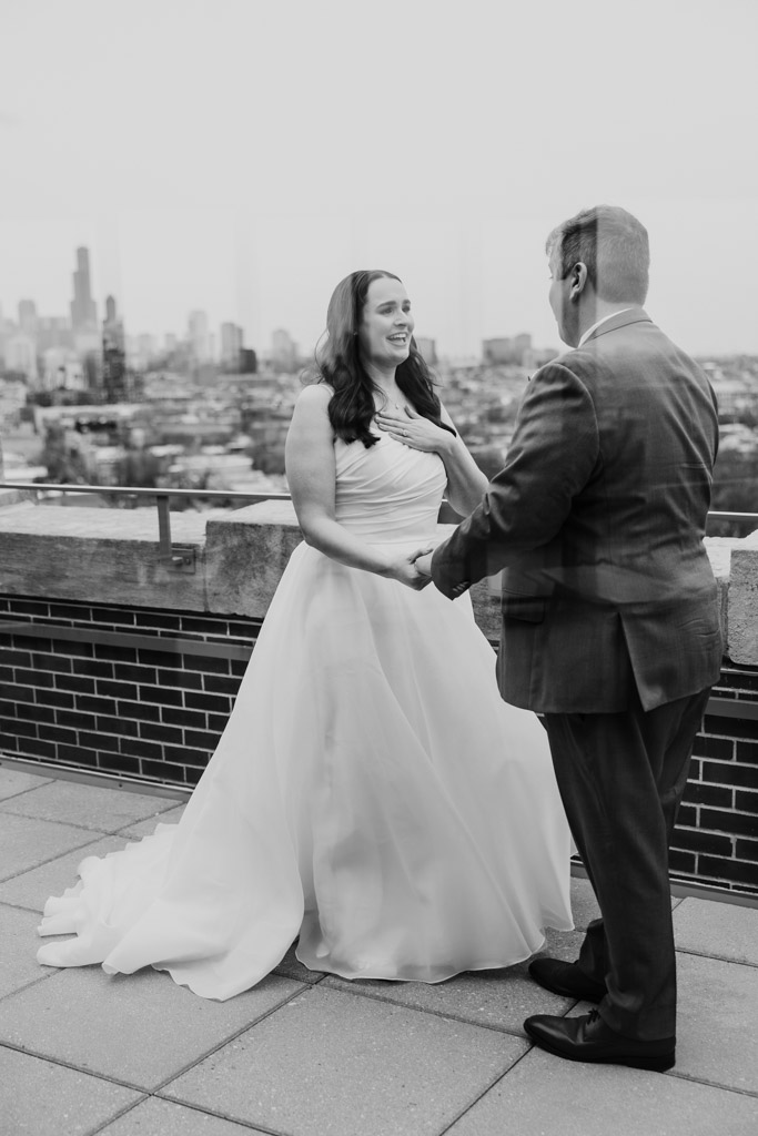 Bride and groom hold hands during their first look on the rooftop of The Robey outside Up Room, the Chicago skyline blurred in the background, black and white photo