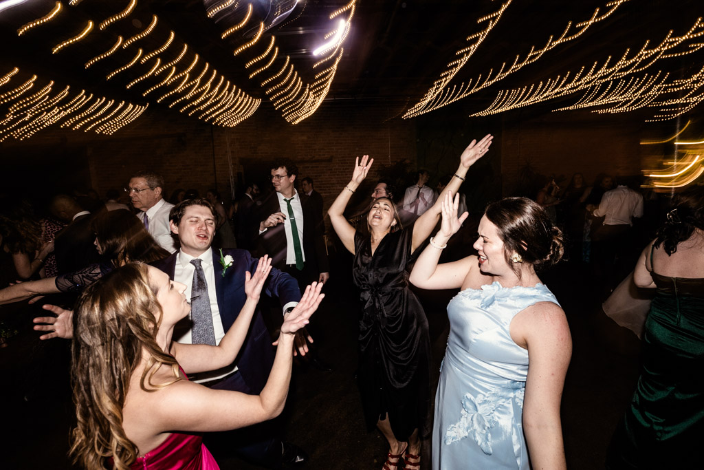 Guests in formal attire dance with raised arms under string lights during wedding reception at The Arbory