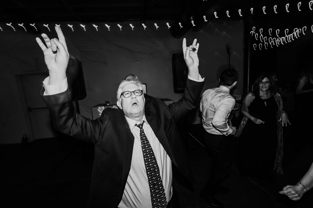 Guest in suit raises hands and dances during wedding reception at The Arbory, while others dance in the background