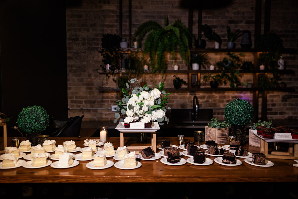 Slices of cake on plates sit on a wooden dessert table at The Arbory, with green plants and white flowers in the background