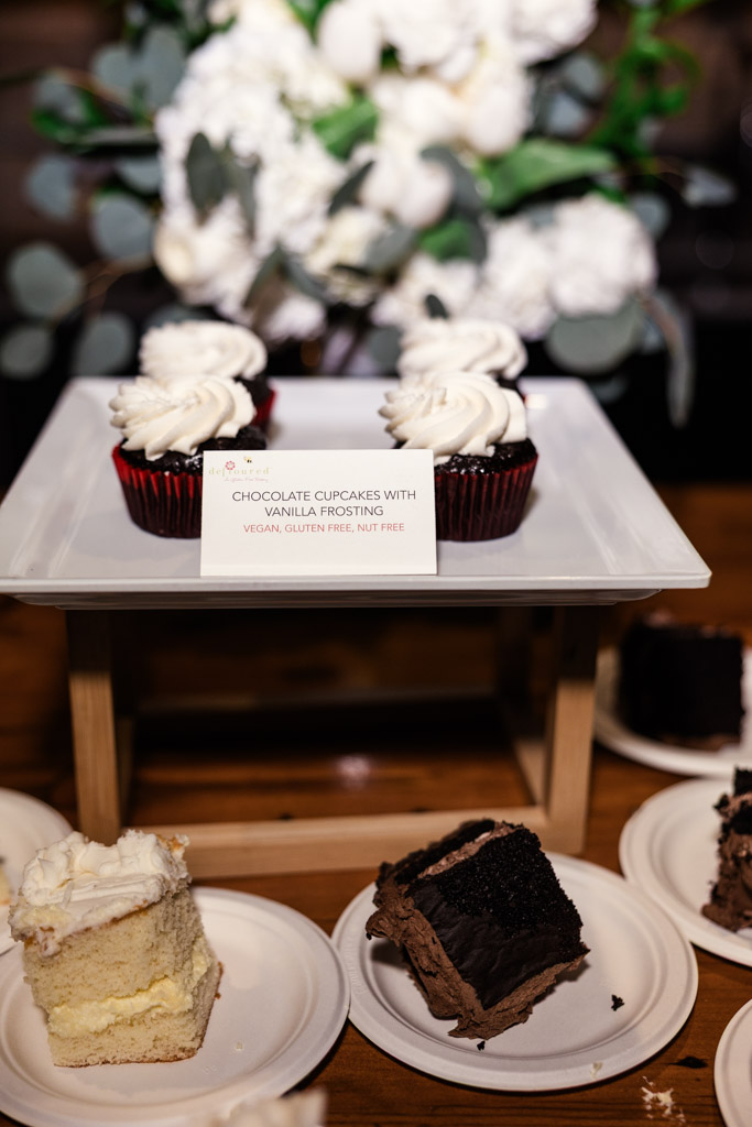 Chocolate cupcakes with vanilla frosting on a tray, accompanied by cake slices on plates in front and delicate flowers in the background fior wedding reception at The Arbory