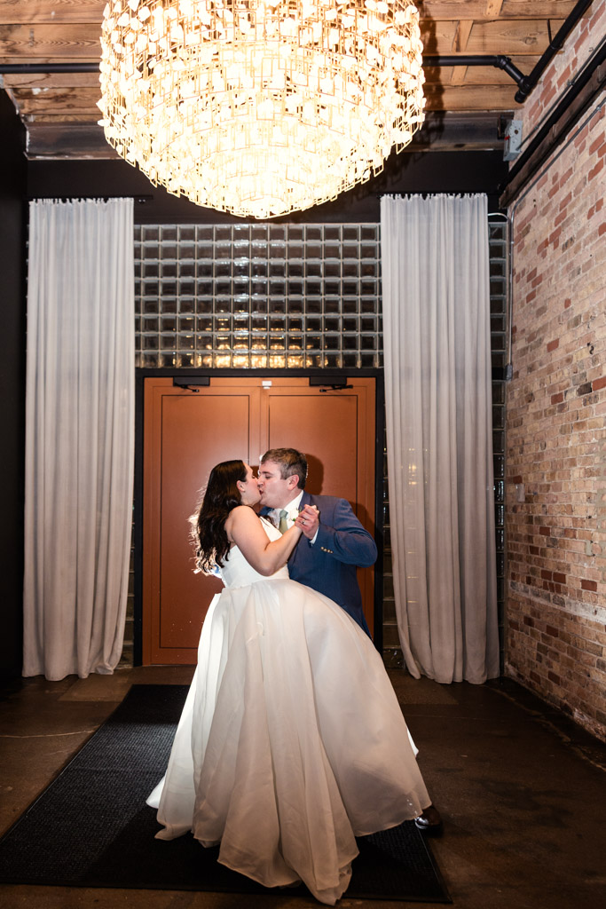 Bride and groom kiss under a large chandelier in the entryway of The Arbory. wedding venue