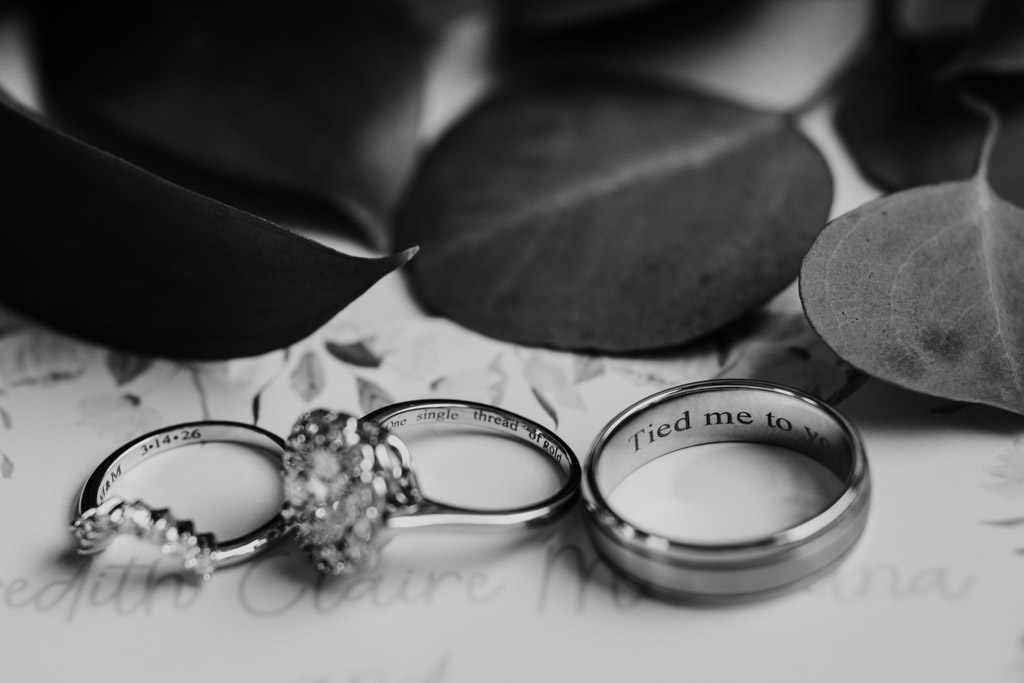 Three rings with engravings rest on paper, surrounded by leaves in a black and white close-up for wedding celebration at The Arbory