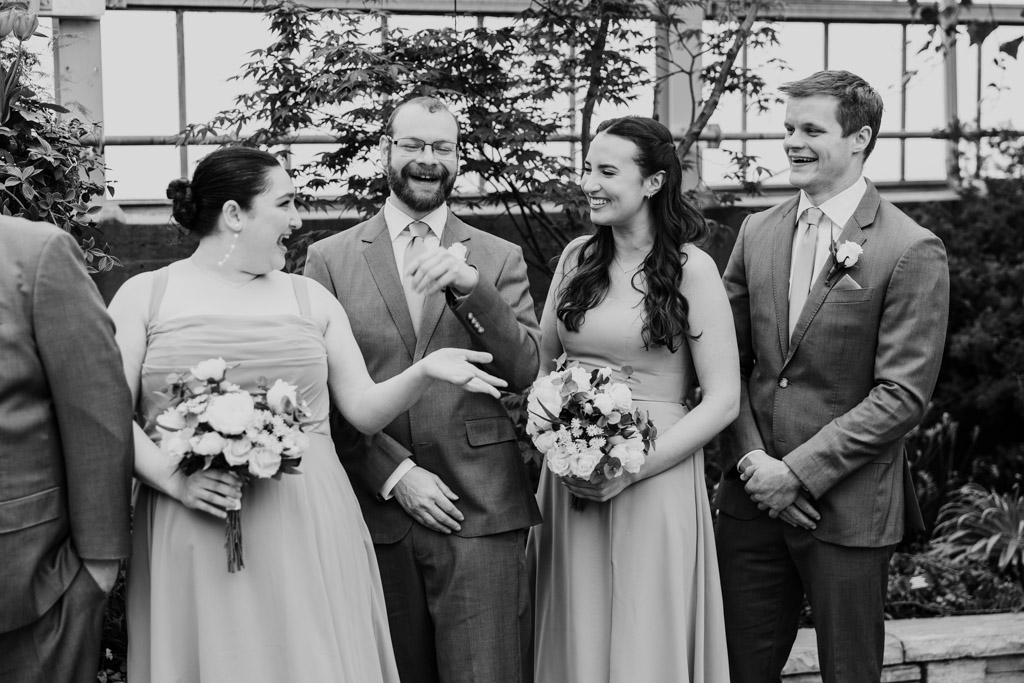 Black and white photo of four members of the wedding party laugh and chat inside Garfield Park Conservatory, two holding bouquets