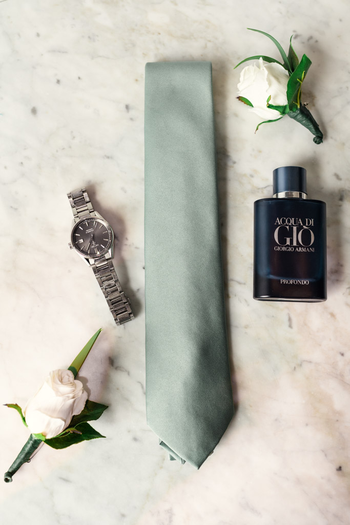 A light green tie, wristwatch, cologne bottle, and two white boutonnieres arranged on a marble surface for wedding at The Arbory in Chicago