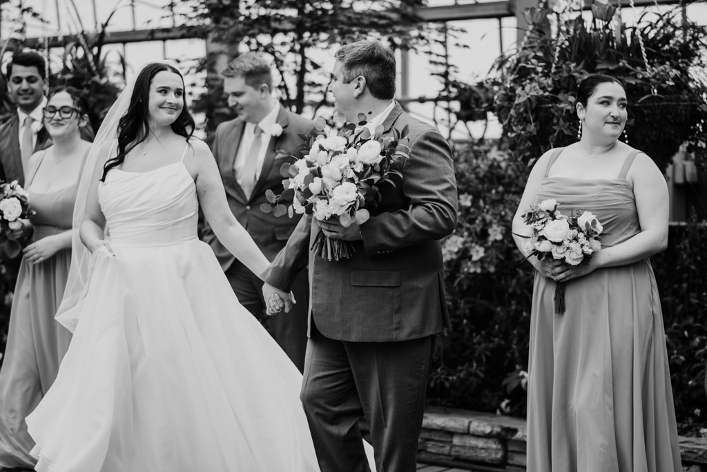 Black adn white photo of bride and groom holding hands, smiling, walking with their wedding party in formal attire through Garfield Park Conservatory