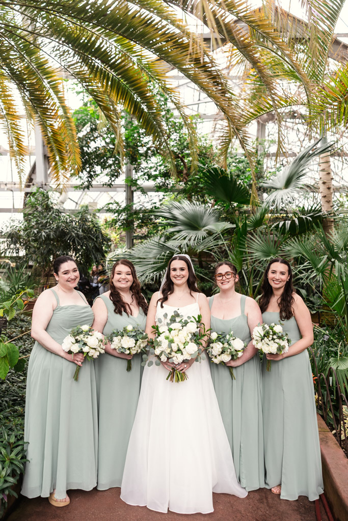 Portrait of bride in white gown and bridesmaids in light green dresses standing together holding bouquets inside Garfield Park Conservatory