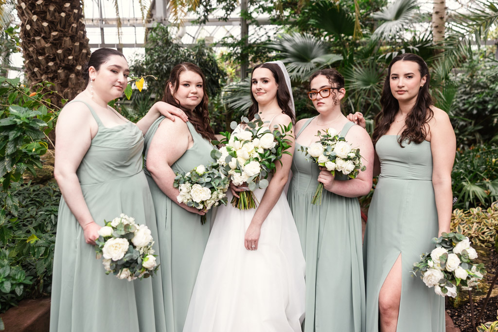 Bride in white dress poses with four bridesmaids in light green gowns at Garfield Park Conservatory, all holding white bouquets