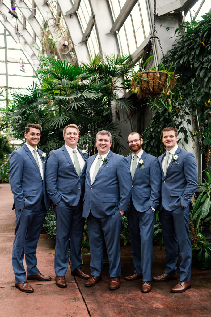 Groom and groomsmen in blue suits pose together, smiling, inside Garfield Park Conservatory filled with lush green plants