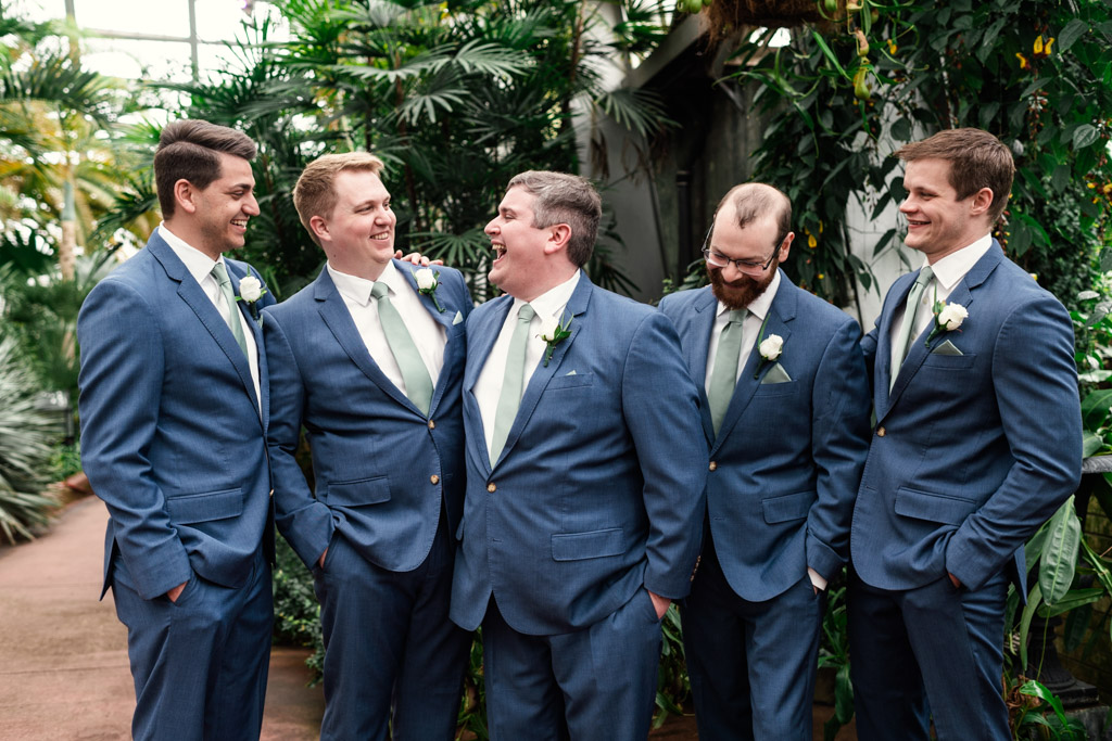 Groom and groomsmenin blue suits with white boutonnieres stand together, smiling and laughing inside Garfield Park Conservatory with lush green plants in the background