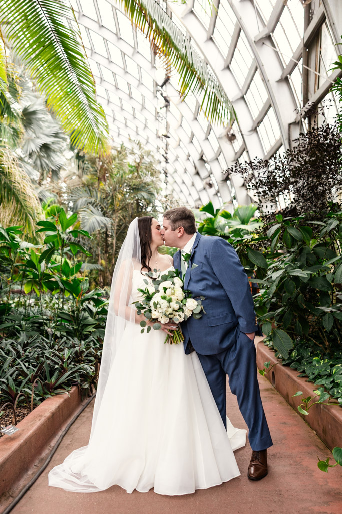 Bride and groom kiss on a path inside Garfield Park Conservatory, bride holding a white bouquet, both dressed in wedding attire