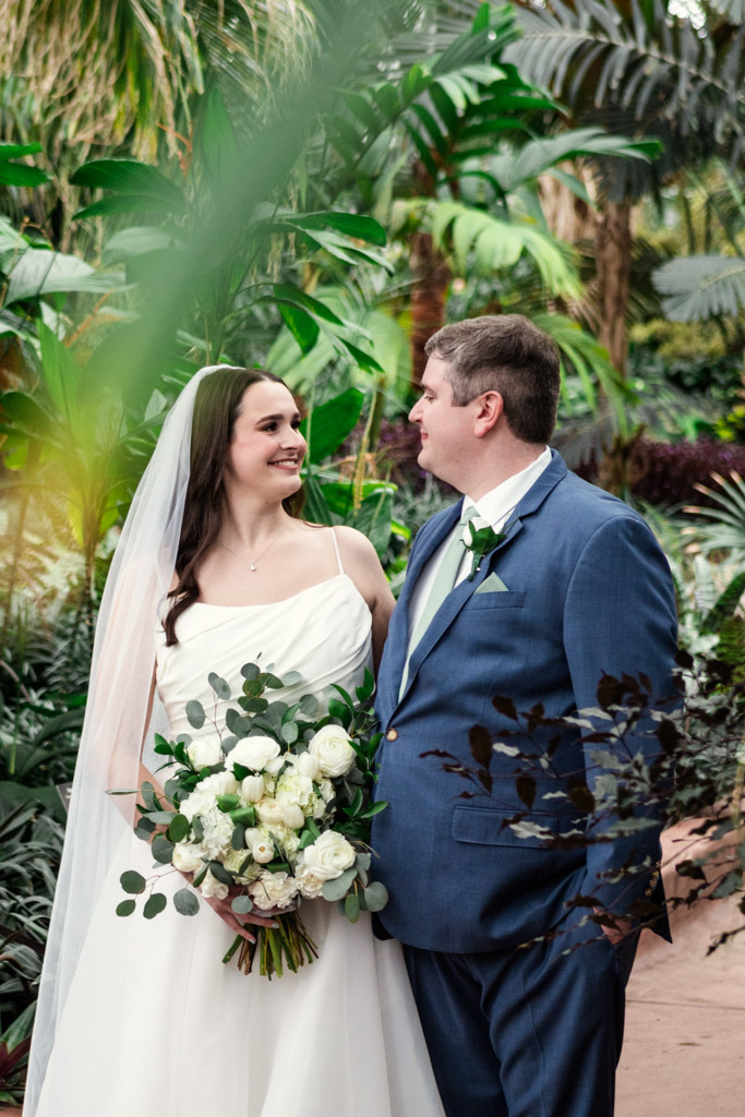 A bride and groom smiling at each other, standing in a lush, green garden with tropical plants at Garfield Park Conservatory