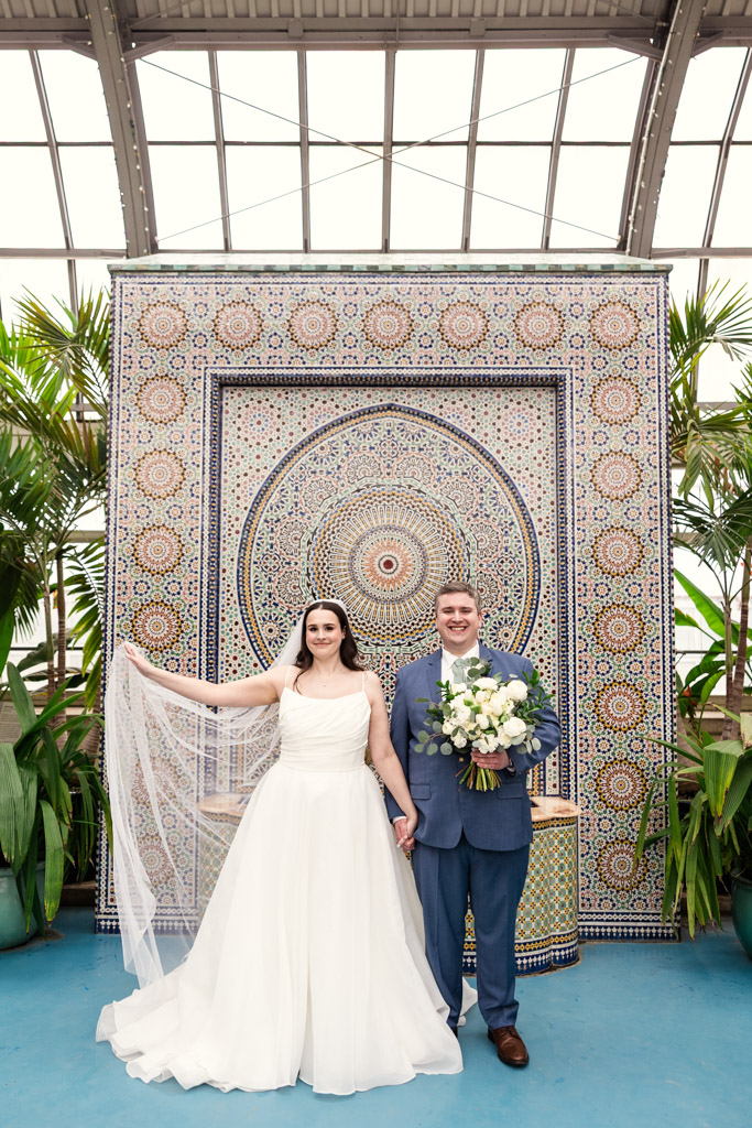 Bride and groom stand smiling in front of a colorful mosaic wall at Garfield Park Conservatory surrounded by lush green plants