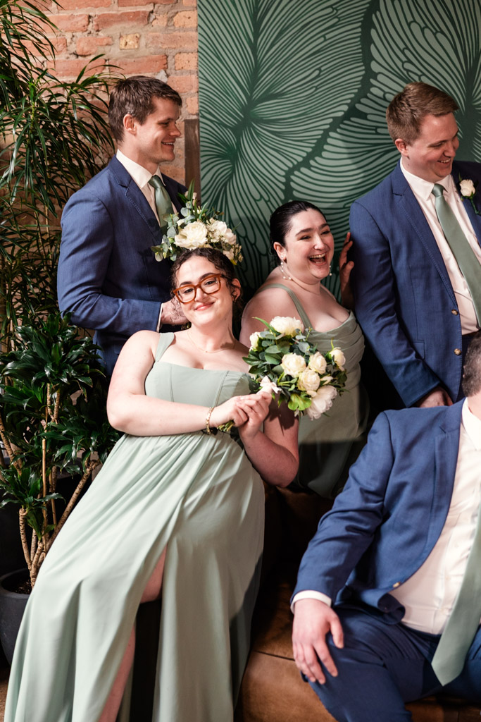 Four weddig party members in green and blue attire smile and pose indoors at The Arbory, holding white flower bouquets, with lush plants behind them