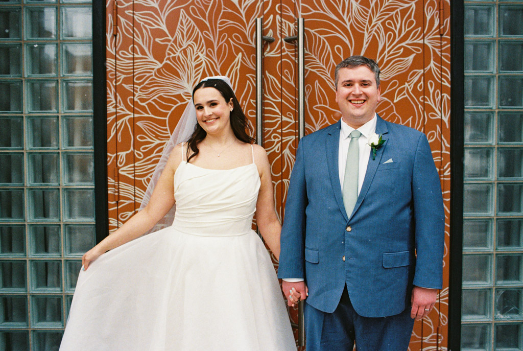 Bride in white dress and groom in blue suit smile, standing in front of ornate orange doors with leaf patterns at The Arbory