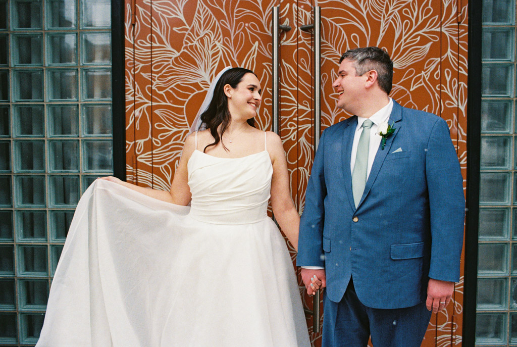 Film photo of bride and groom smiling at each other, holding hands in front of a decorative orange door at The Arbory