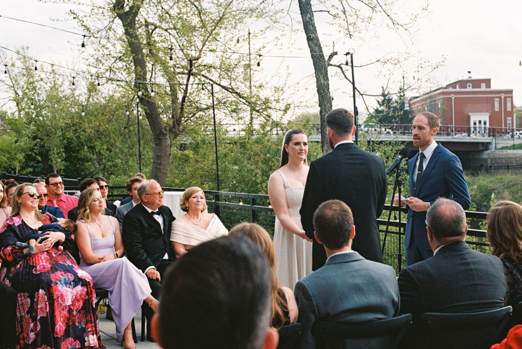 Film photo of outdoor wedding ceremony at River Room with bride and groom holding hands, guests seated, and an officiant speaking