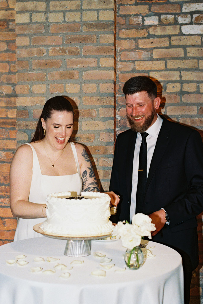 Film photo of happy newlywed couple cutting a white cake together at a table decorated with white flowers and petals inside River Room