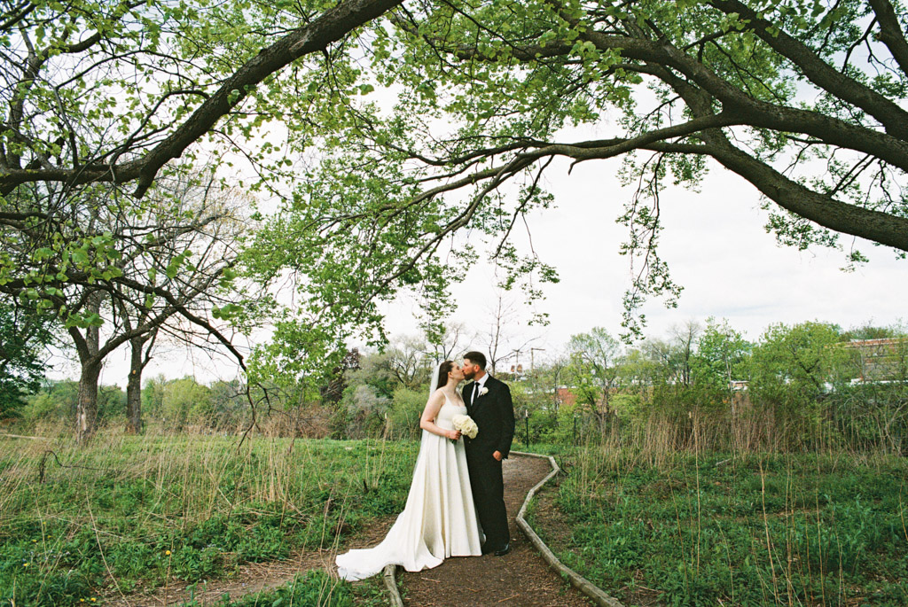 Film photo of bride and groom kissing on a narrow path in a green, wooded outdoor setting under tree branches in Horner Park