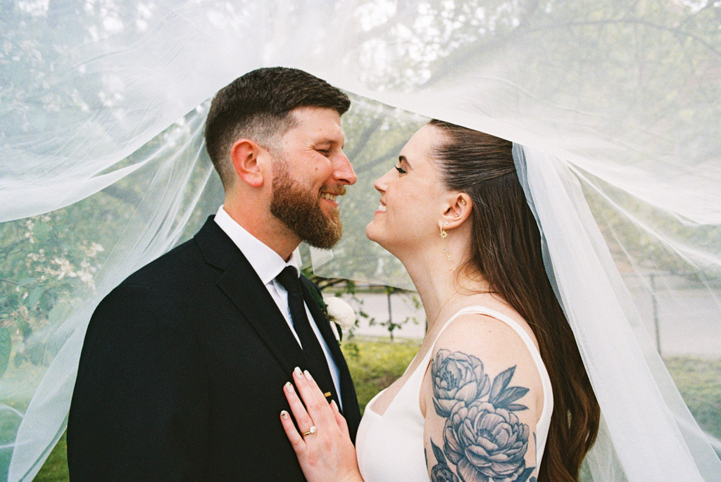 Film photo of bride and groom smiling under a veil outdoors in Horner Park, facing each other; bride has floral tattoo