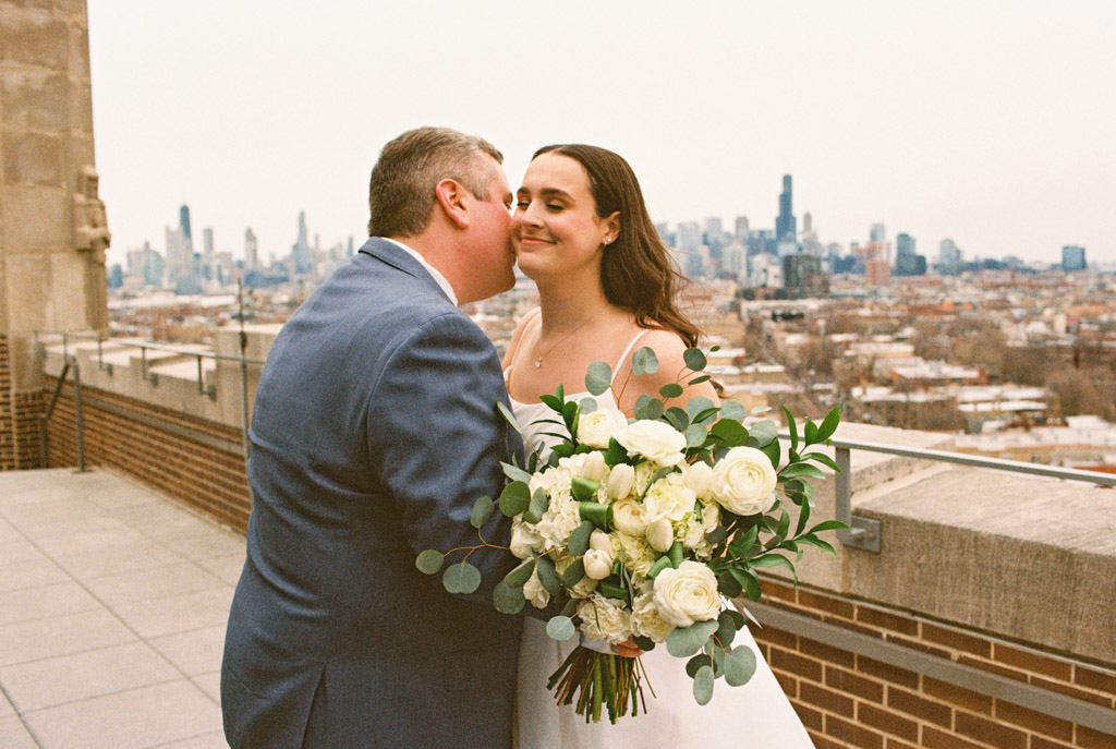 Film photo of groom kissing his bride’s cheek on the rooftop of The Robey, with the Chicago skyline in the background