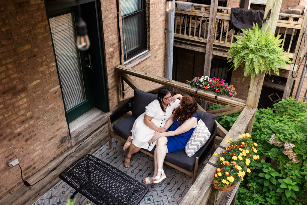 Two women sit closely on a couch on their balcony deck, surrounded by flowers during their Wicker Park engagement session