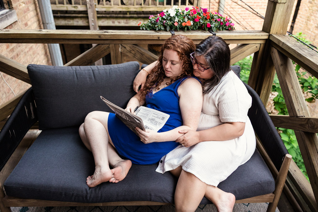 Engaged couple sits closely on an outdoor couch on their balcony in Wicker Park, doing a crossword puzzle together, with flowers on the railing behind them